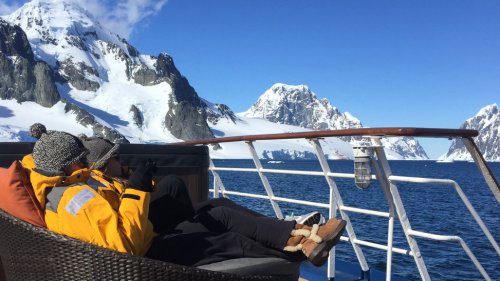Two people relax on a boat, surrounded by snowy mountains and clear blue water under a bright sky.