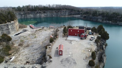 Aerial view of a quarry with a red building, water, and rocky cliffs surrounding a calm, blue lake.