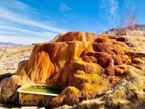 Colorful mineral formations rise from the ground, with a small green pool at the base under a clear blue sky.
