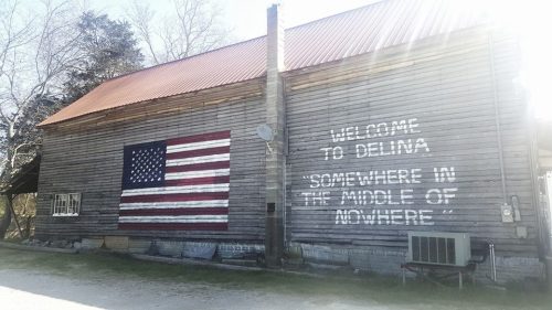 A rustic wooden building with an American flag mural and the text "Welcome to Delina, somewhere in the middle of nowhere."