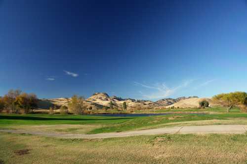 A serene landscape featuring a calm pond, rolling hills, and a clear blue sky. Green grass in the foreground.