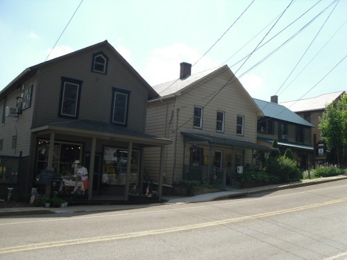 A quaint street scene featuring two houses and a shop, with power lines overhead and greenery along the sidewalk.