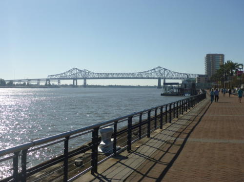 A sunny riverside view featuring a bridge, waterfront walkway, and palm trees along the riverbank.
