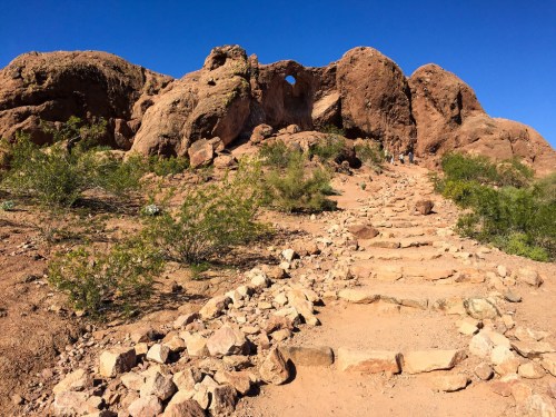 A rocky trail leads up a desert landscape with large boulders and a clear blue sky in the background.