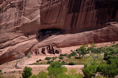 A rocky cliff with ancient structures nestled in a canyon, surrounded by green vegetation and trees.
