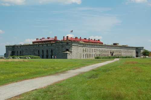 Historic stone fort with a red roof, surrounded by green grass and a pathway, under a blue sky with clouds.