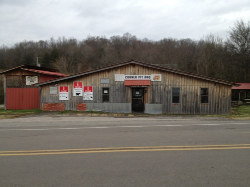 A rustic wooden building with a sign reading "Corner Pit BBQ," surrounded by trees and a rural road.