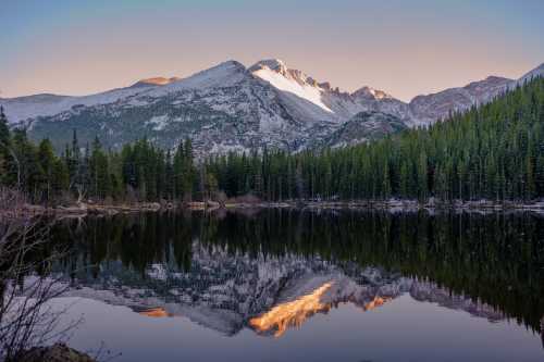 A serene mountain landscape with snow-capped peaks reflecting in a calm lake, surrounded by evergreen trees.