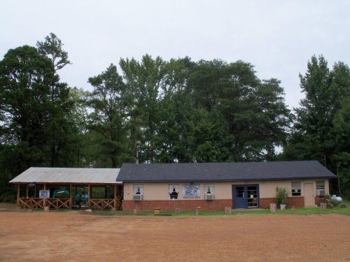 A single-story building with a porch, surrounded by trees and gravel, under a cloudy sky.