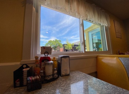 A view from inside a diner, showing a table with condiments and a window revealing a bright, sunny day outside.
