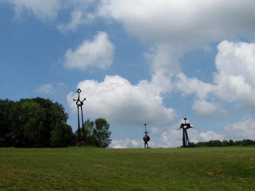 Three large metal sculptures stand on a grassy hill under a partly cloudy sky.