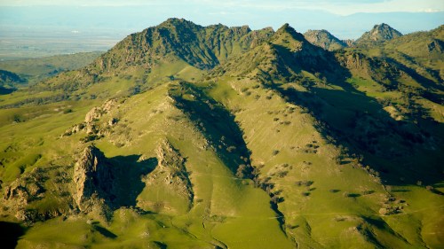 Aerial view of lush green mountains with rocky peaks under a clear blue sky.