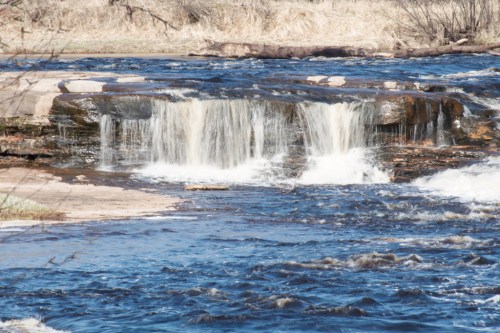 A serene waterfall cascading over rocks into a flowing river, surrounded by grassy banks and bare trees.