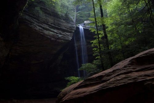 A serene waterfall cascades down rocky cliffs, surrounded by lush green trees in a tranquil forest setting.