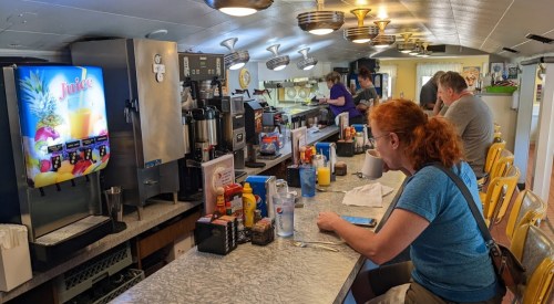 A diner scene with a woman sitting at the counter, drinks and condiments visible, and staff serving in the background.