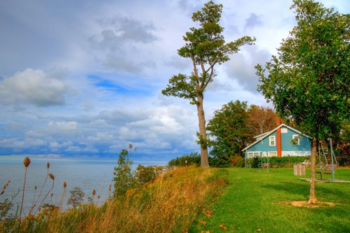 A scenic view of a house by the water, surrounded by trees and grassy land under a cloudy sky.