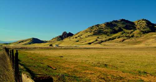 A wide, open landscape featuring rolling hills, a clear blue sky, and a wooden fence in the foreground.