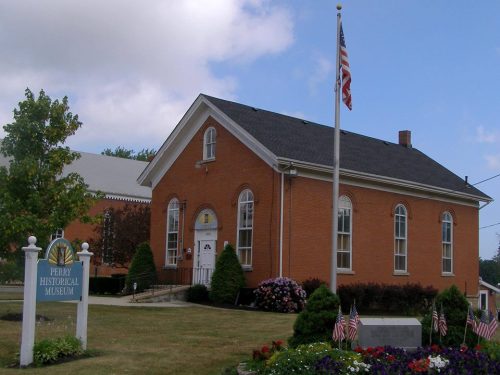 A brick building with arched windows, the Perry Historical Museum, surrounded by flowers and an American flag.