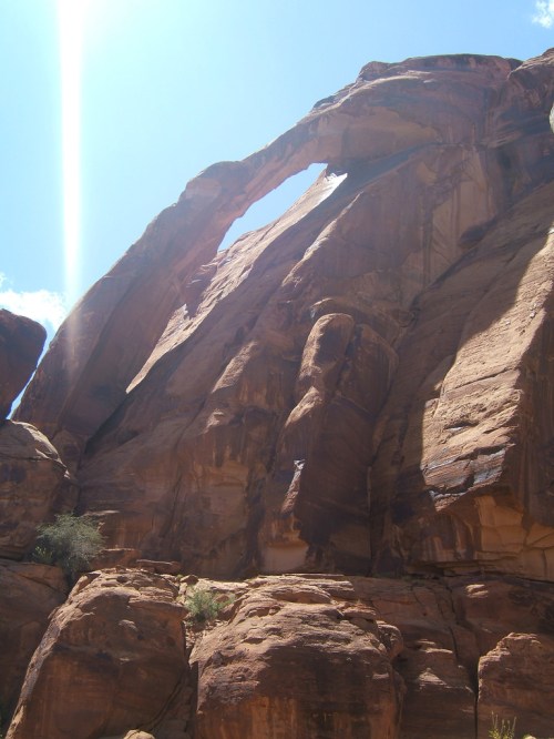 A towering rock formation with a natural arch under a bright blue sky, surrounded by rugged terrain.
