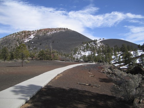 A winding path leads through a volcanic landscape with hills, trees, and a blue sky with scattered clouds.