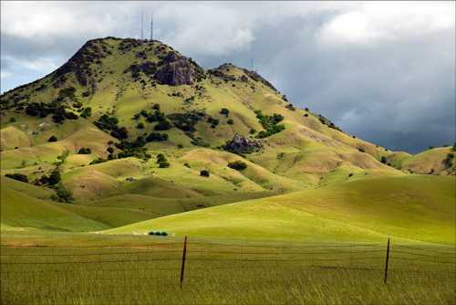 Lush green hills under a cloudy sky, with a mountain peak featuring communication towers in the background.