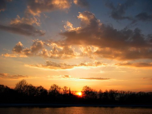 A serene sunset over a calm lake, with vibrant clouds and silhouetted trees along the horizon.
