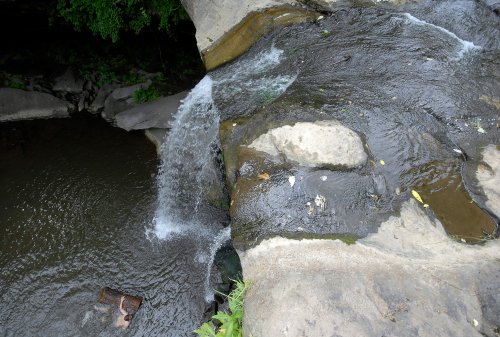 A small waterfall cascades over rocks into a dark stream, surrounded by greenery and scattered debris.