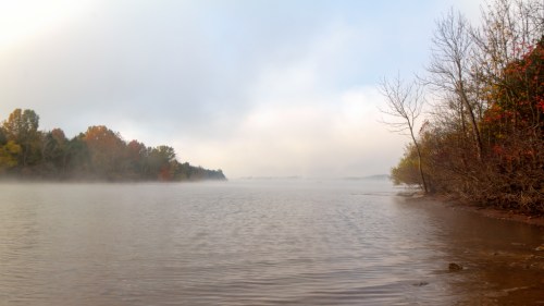 A serene lake scene with fog, surrounded by trees displaying autumn colors under a soft, misty sky.