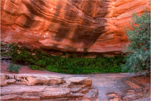 A vibrant red rock formation with lush green vegetation at the base, set in a serene natural landscape.