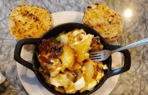 A cast iron skillet filled with scrambled eggs, vegetables, and potatoes, served with two toasted bread slices.