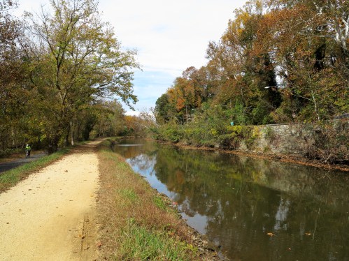 A peaceful canal lined with trees in autumn colors, with a path beside it and a cyclist in the distance.