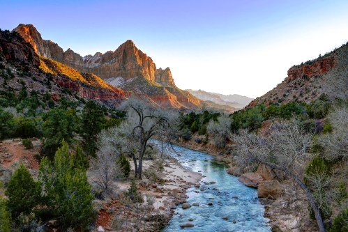 A serene river flows through a canyon, surrounded by towering red rock formations and sparse trees under a clear sky.