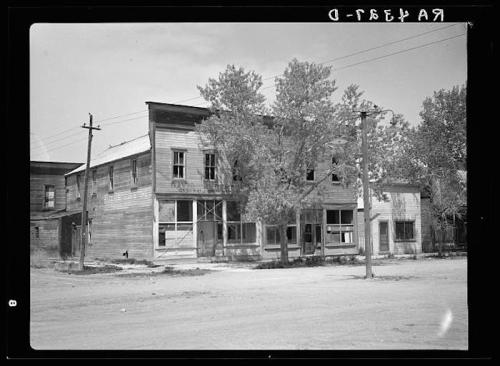 Abandoned wooden building with boarded windows, surrounded by trees and power lines, on a dirt road.