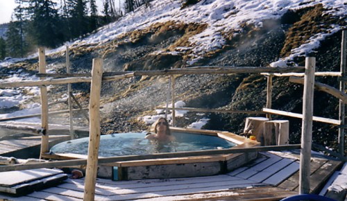 A person relaxes in a hot tub surrounded by snow-covered ground and trees, steam rising in the chilly air.