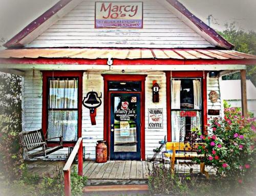 A quaint bakery with a red roof, white siding, and a welcoming porch adorned with flowers and a sign for Marcy Jo's.