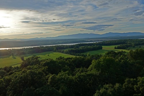 A scenic view of lush green fields and a river, with mountains and a cloudy sky in the background.