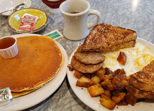 A breakfast spread featuring pancakes, eggs, sausage, toast, and home fries, with coffee and syrup on the side.
