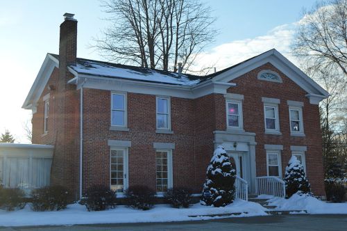 A brick house with a white porch, surrounded by snow and trees, under a clear blue sky.