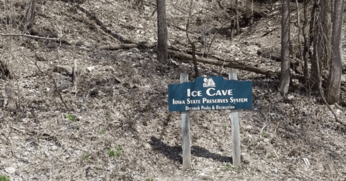 Sign for Ice Cave, Iowa State Preserves System, surrounded by a wooded area with bare trees and rocky terrain.