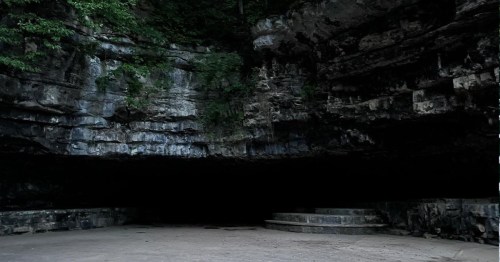 A dark cave entrance surrounded by rocky walls and greenery, with stone steps leading into the cave.