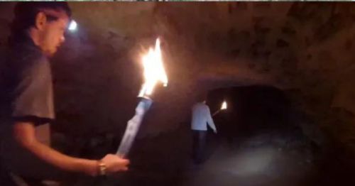 Two people walking in a dark cave, holding torches that illuminate the rocky surroundings.