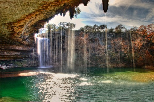 A serene waterfall cascades into a green pool, surrounded by rocky cliffs and trees under a blue sky.