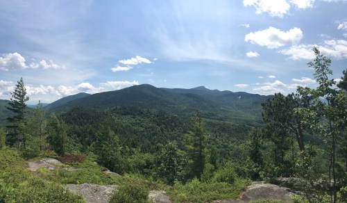 A panoramic view of lush green mountains under a bright blue sky with scattered clouds.
