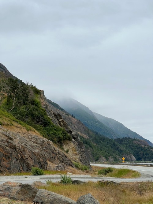 A misty mountain landscape with lush greenery and rocky terrain, alongside a winding road under a cloudy sky.
