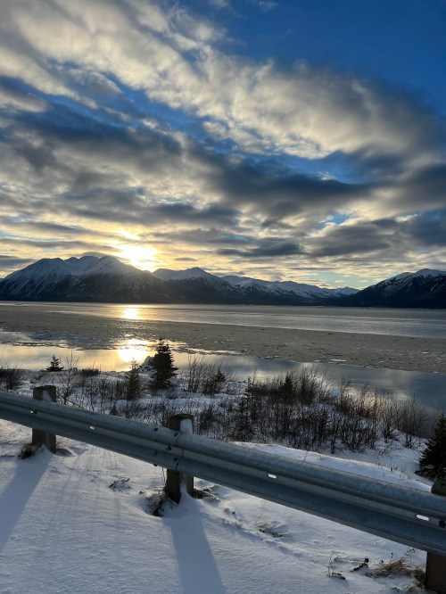 A scenic view of a lake surrounded by mountains, with a sunset reflecting on the water and snow-covered ground.