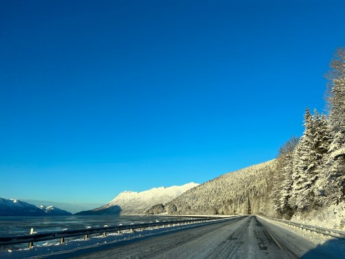 A snowy road winds along a frozen lake, flanked by snow-covered mountains under a clear blue sky.