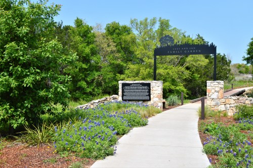 Pathway leading to a family garden entrance, framed by greenery and stone structures under a clear blue sky.