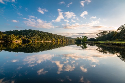 A serene lake reflects a blue sky and clouds, surrounded by lush green hills and trees.
