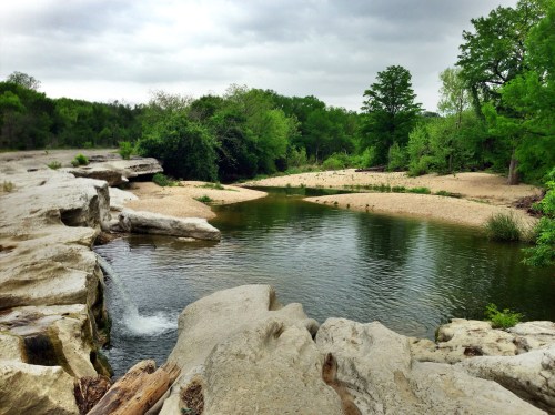 A serene river scene with rocky edges, sandy banks, and lush greenery under a cloudy sky.