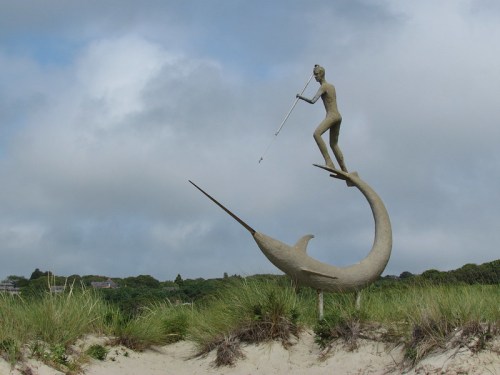 A sculpture of a figure playing a flute while balancing on a large fish, set against a cloudy sky and grassy landscape.